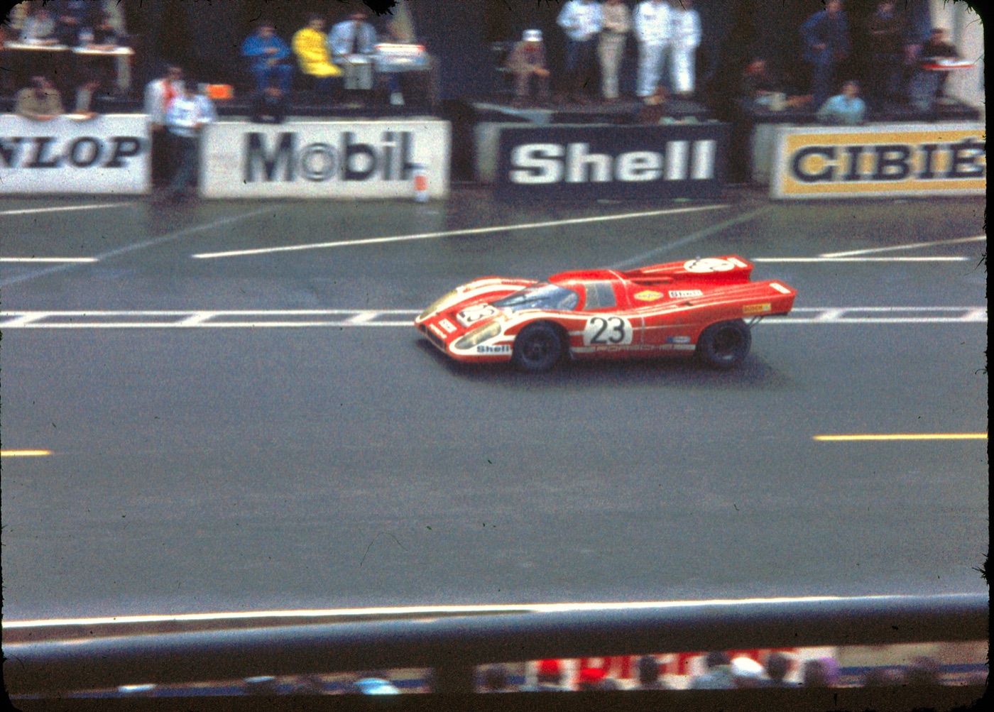 Red and white Porsche 917K number 23 on a wet track in front of Mobil, Shell and Cibié advertising boards