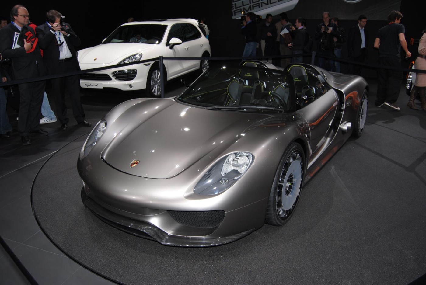 Silver-bronze Porsche 918 Spyder concept roadster on a turntable, photographers behind and a white Cayenne S Hybrid on the left