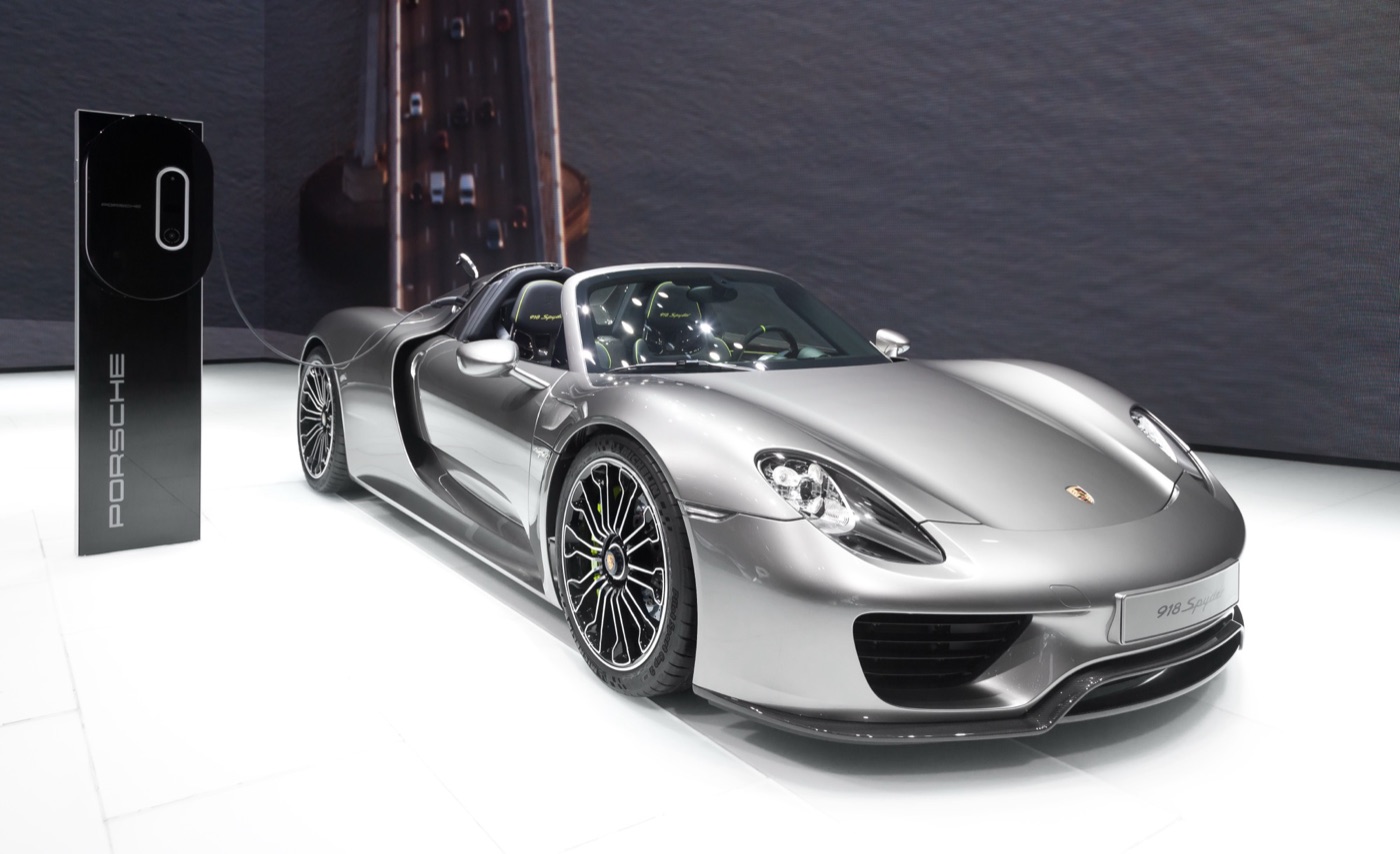 Silver Porsche 918 Spyder roadster on a white show floor, plugged into a Porsche charging column