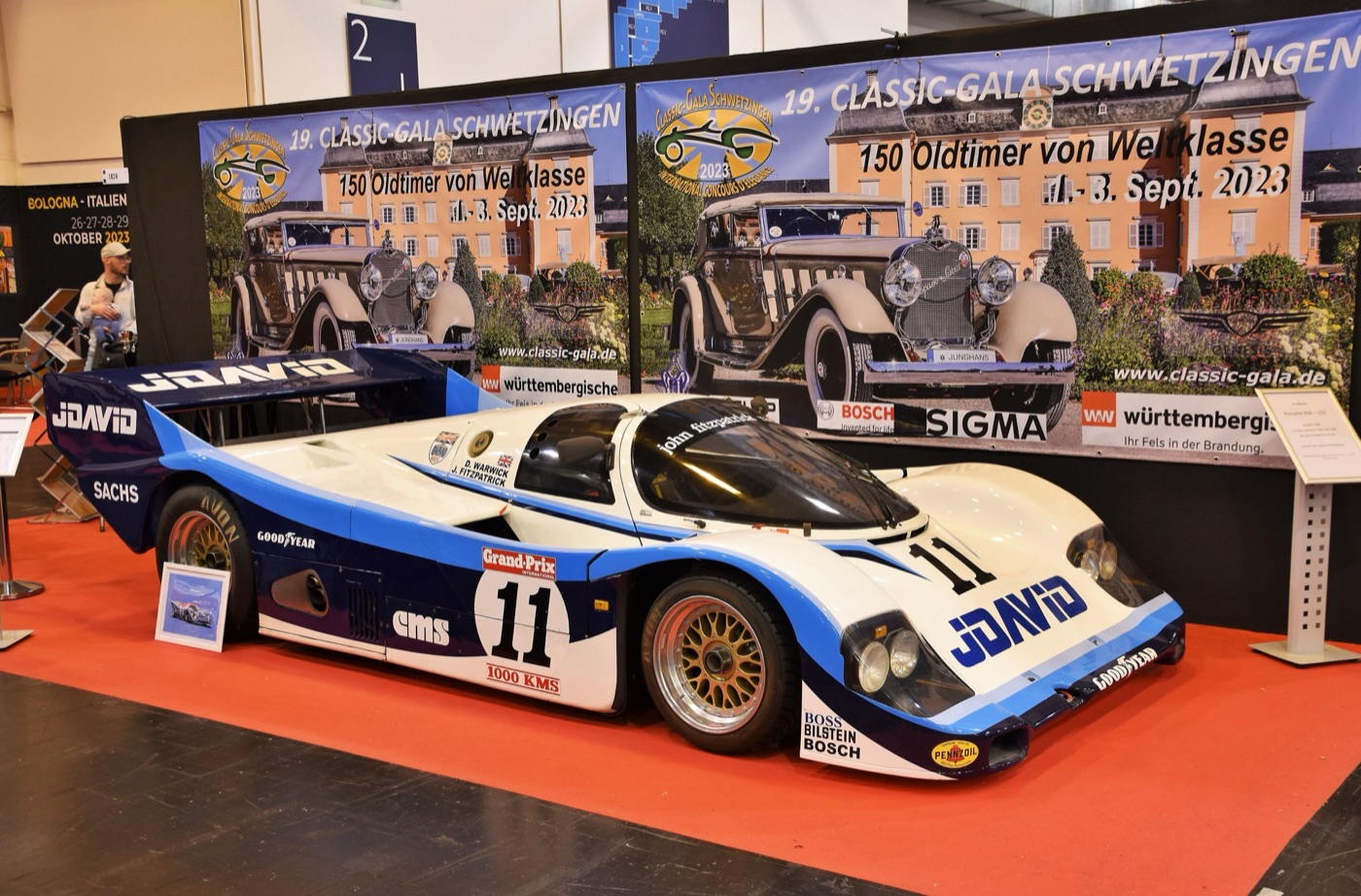 White and blue JDavid-liveried Porsche 956 number 11 on a red carpet at an indoor classic car show