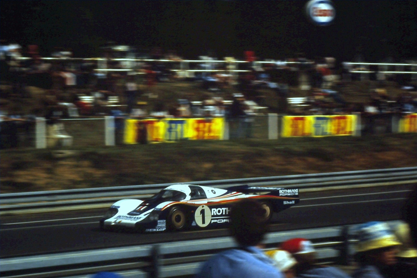 Porsche 956 number 1 in Rothmans colours passing a crowded grandstand behind a guard rail, spectators in the foreground