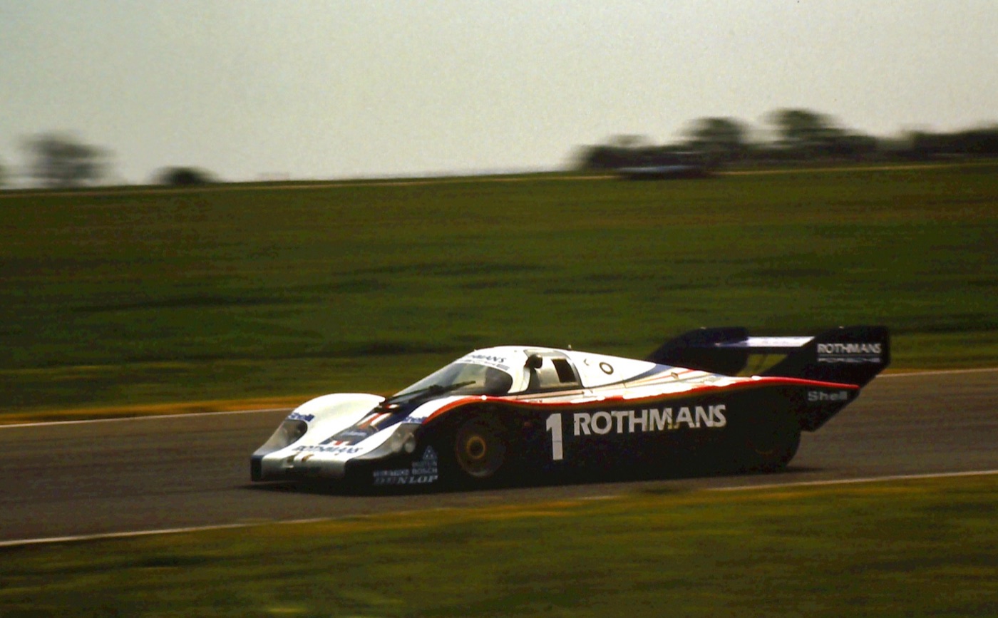 White and blue Porsche 956 number 1 in Rothmans colours cornering on a flat circuit edged with grass