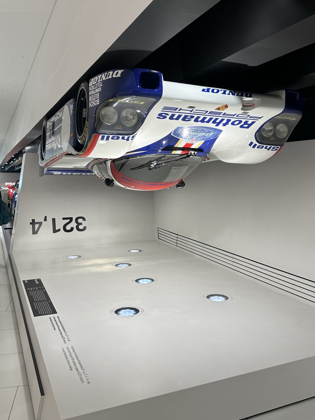 Porsche 956 in Rothmans colours mounted upside down on the ceiling of a museum display, with 321,4 painted on the wall beneath
