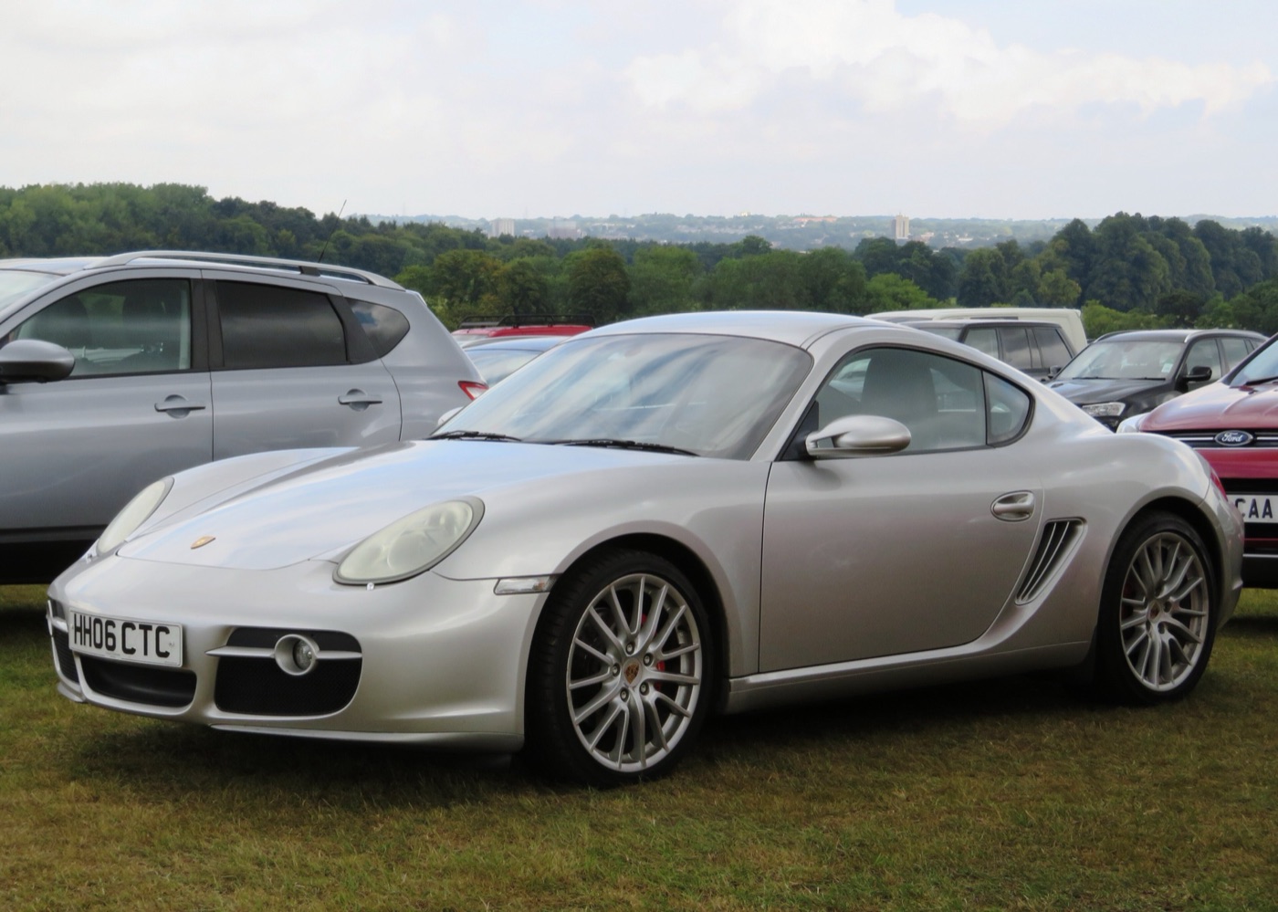 Silver first-generation Porsche Cayman S of 2006, the 987, parked on grass among other cars