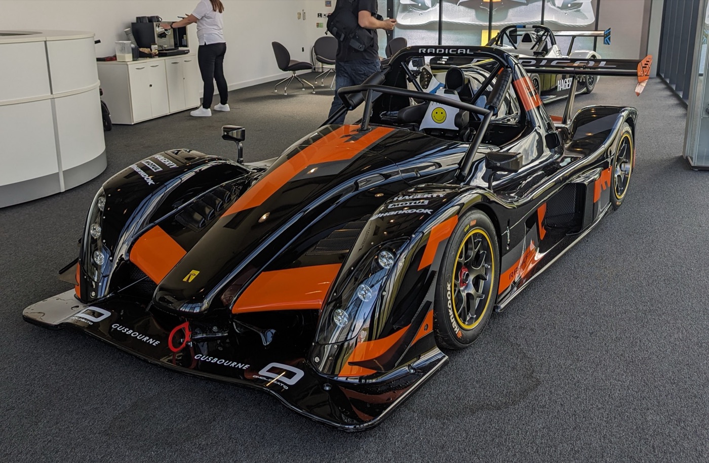 Black and orange Radical SR10 XXR sports-racer with a roll hoop and twin headlamps, parked in a showroom