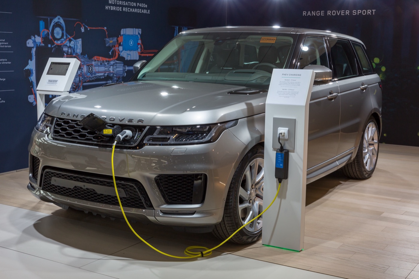 Silver facelifted Range Rover Sport P400e on a motor show stand, a yellow charging cable plugged in behind the grille badge