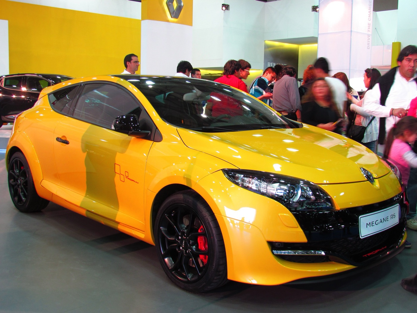 Yellow Megane RS with black wheels, red calipers and a Trophy door graphic on a motor show stand