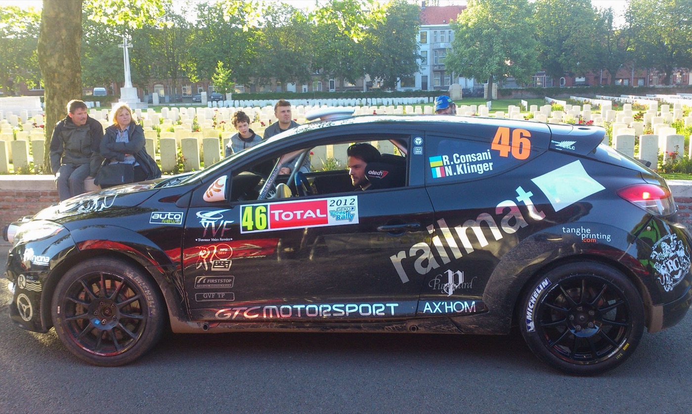 Black Megane RS N4 rally car with sponsor decals and number 46, a driver inside, parked beside a war cemetery