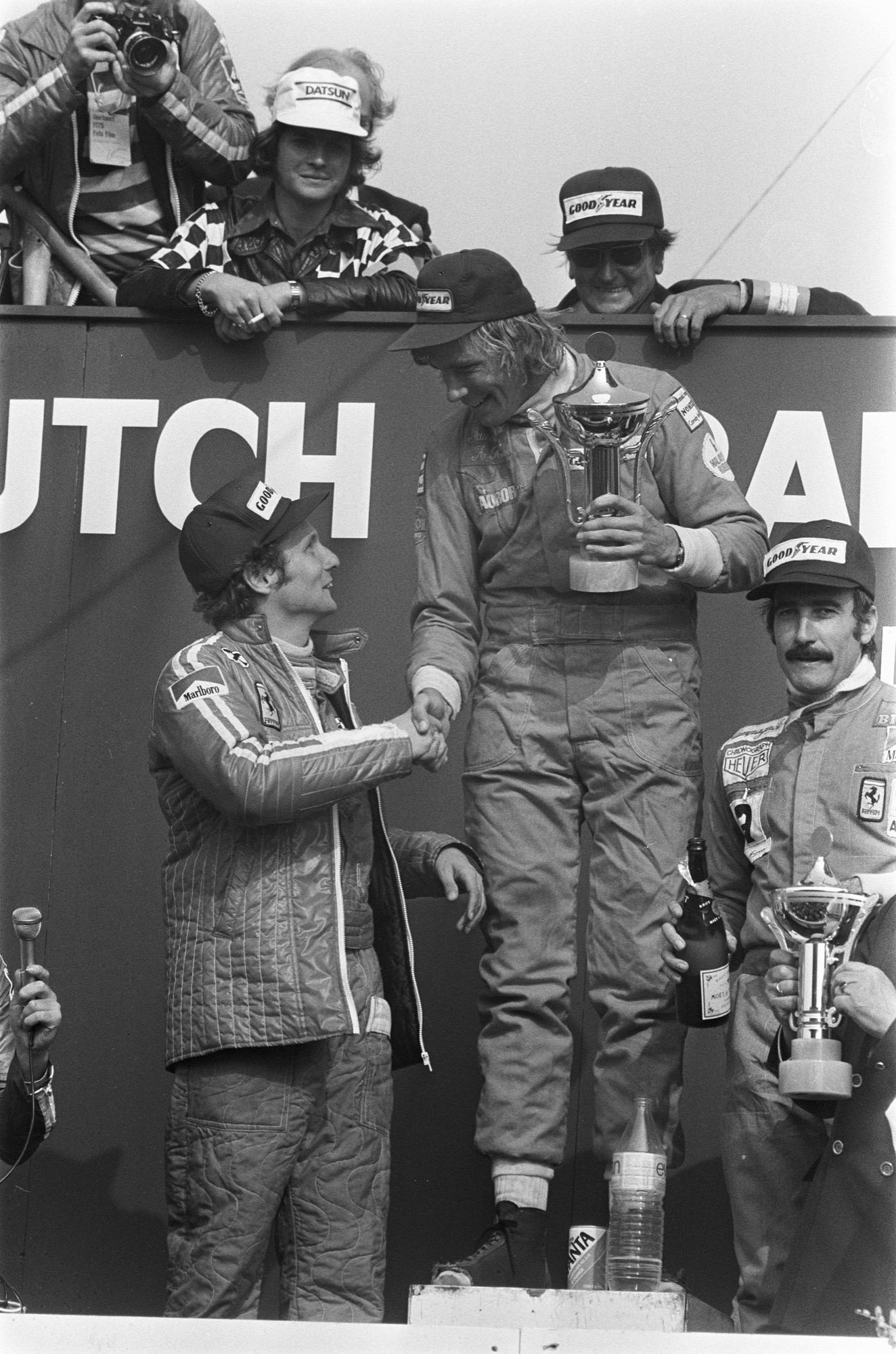 James Hunt on the top step of the podium holding a trophy and shaking hands with Niki Lauda in a Ferrari jacket, Clay Regazzoni beside them