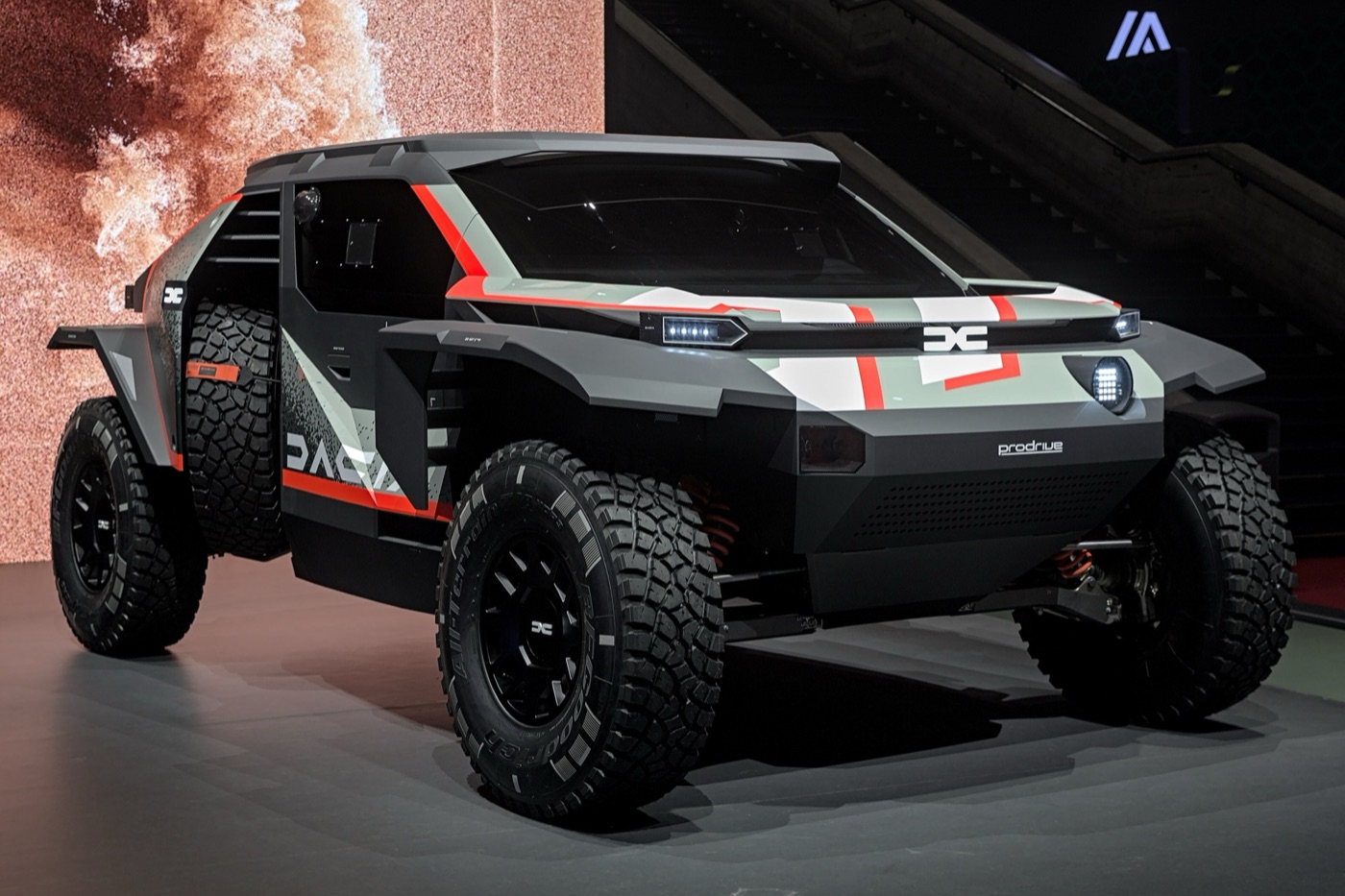 Grey Dacia Sandrider rally-raid prototype with red stripes and large off-road tyres on a show stand