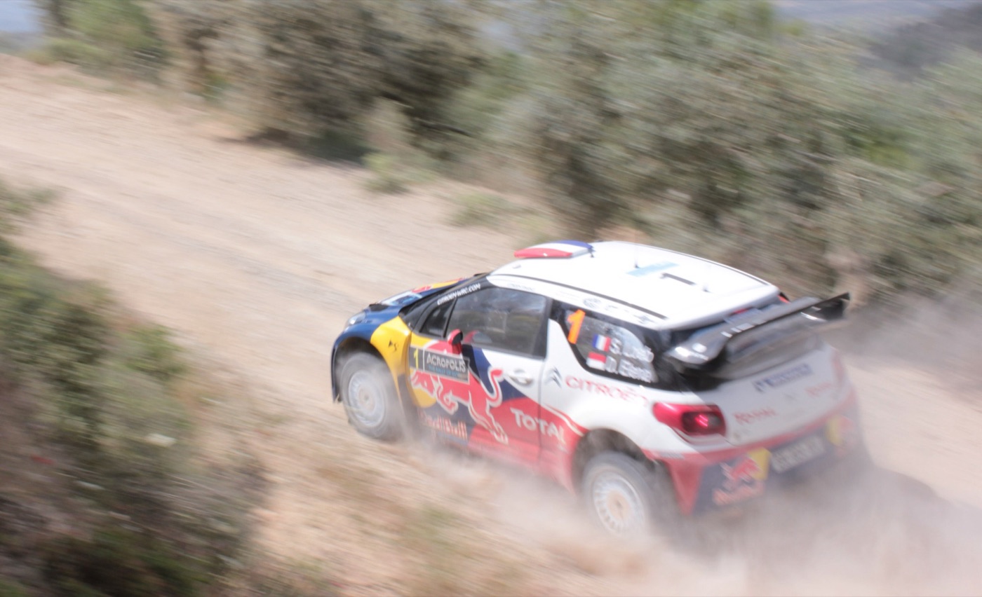 Loeb’s white, red and blue Citroën DS3 WRC sliding through a dusty gravel corner between bushes