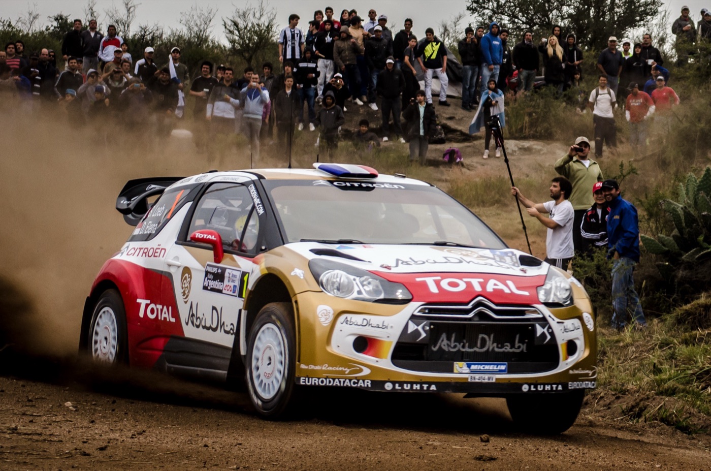 Sébastien Loeb’s gold, white and red Citroën DS3 WRC, sliding on gravel past spectators in Argentina