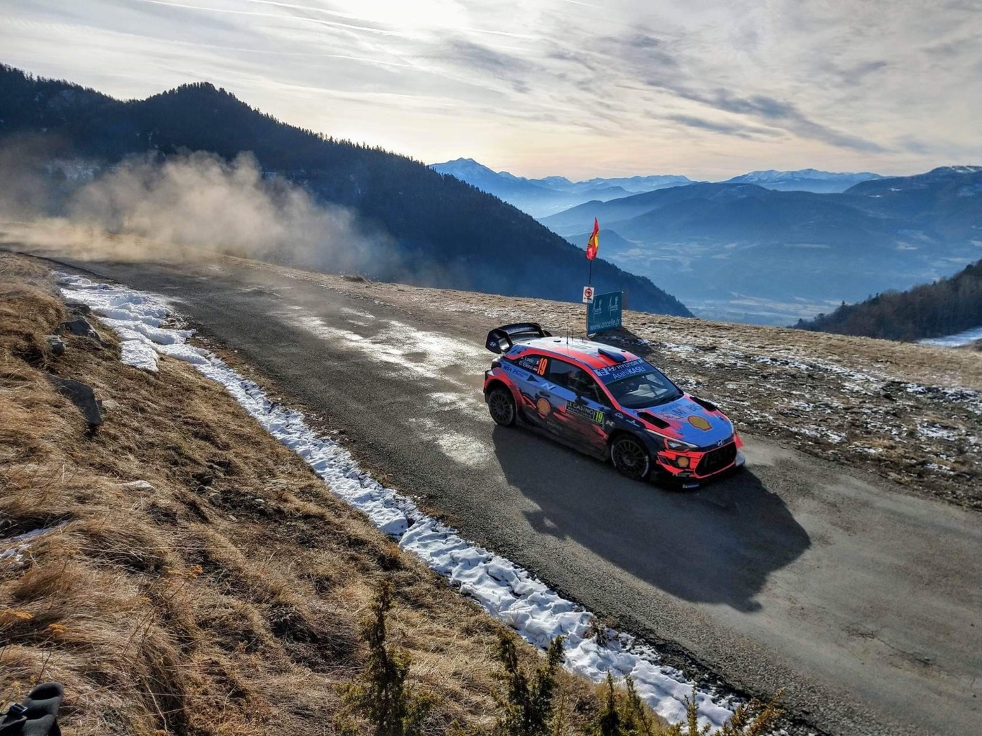 Hyundai i20 Coupe WRC in red, blue and orange livery on a mountain road edged with snow, with Alpine peaks behind