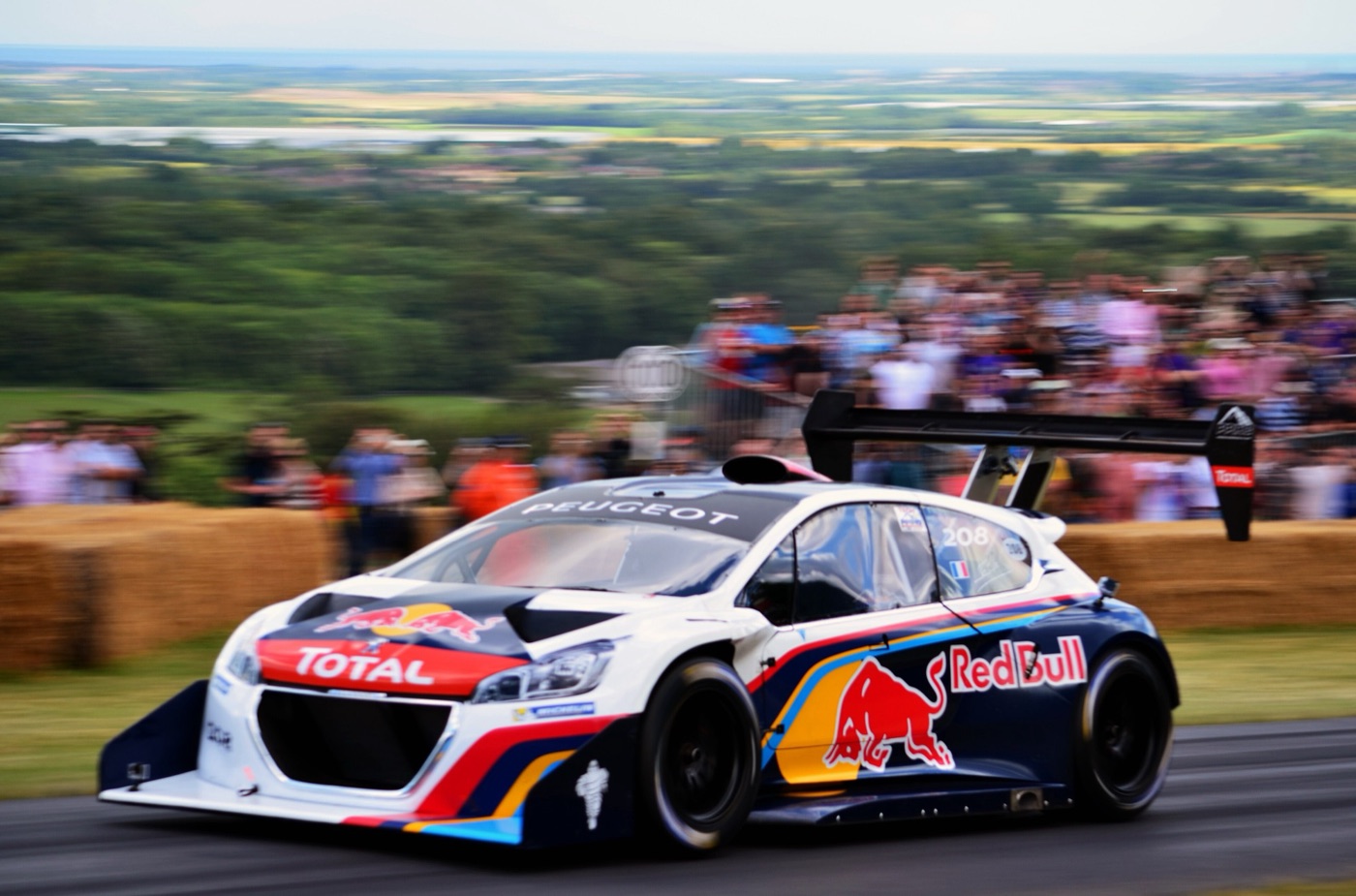 Peugeot 208 T16 Pikes Peak in Red Bull and Total livery with a huge rear wing, passing hay bales and a crowd