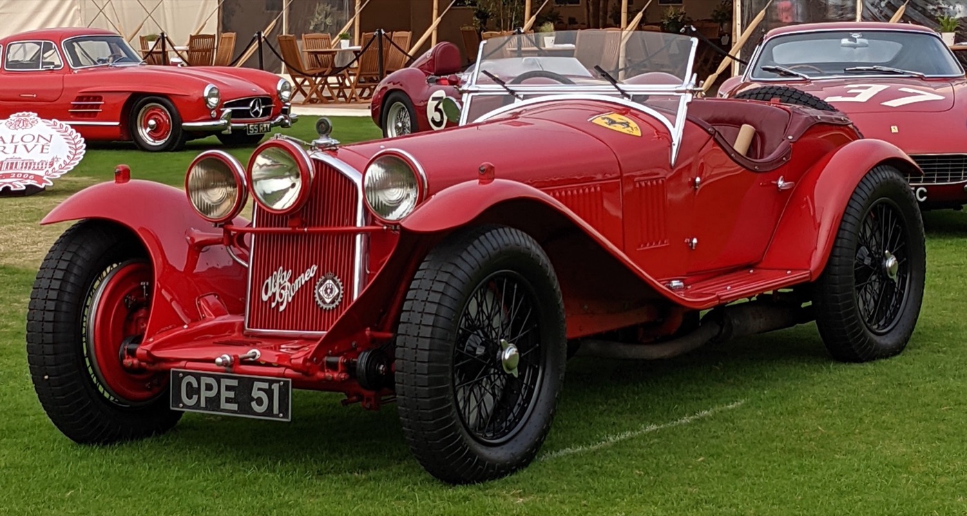 A red 1931 Alfa Romeo 8C 2300 Monza spider with a Scuderia Ferrari badge on its bonnet, a red Mercedes-Benz 300 SL coupé behind and a Salon Privé sign on the left