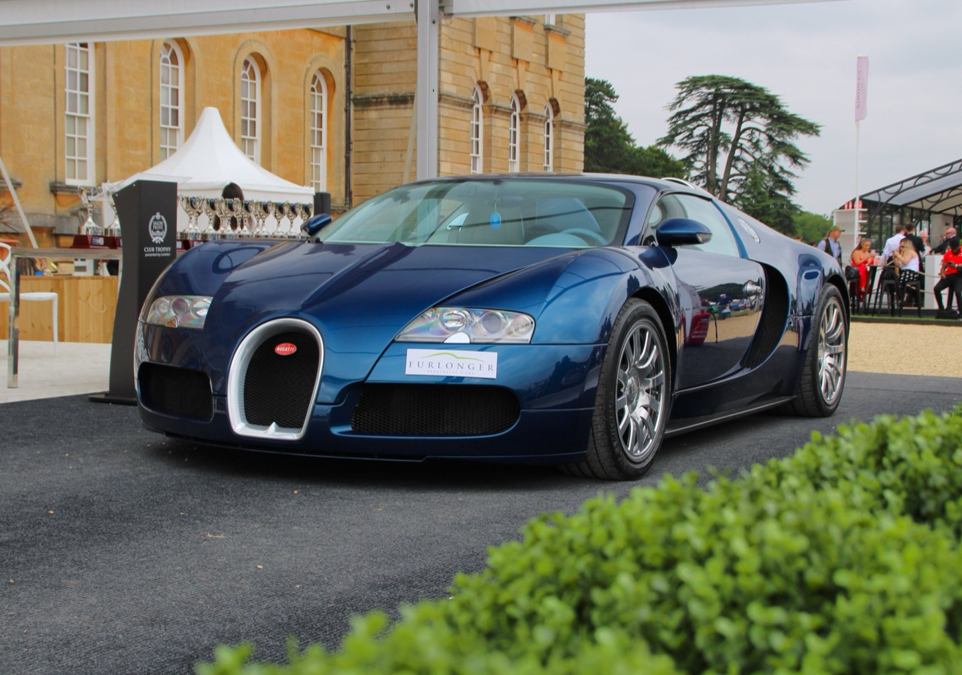 A dark blue Bugatti Veyron on a carpeted stand beside the Club Trophy lectern and a row of silver cups, Blenheim Palace behind