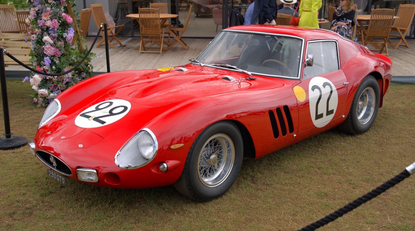 A red Ferrari 250 GTO with race number 22 roped off on the grass in front of a hospitality pavilion, beside a column of flowers