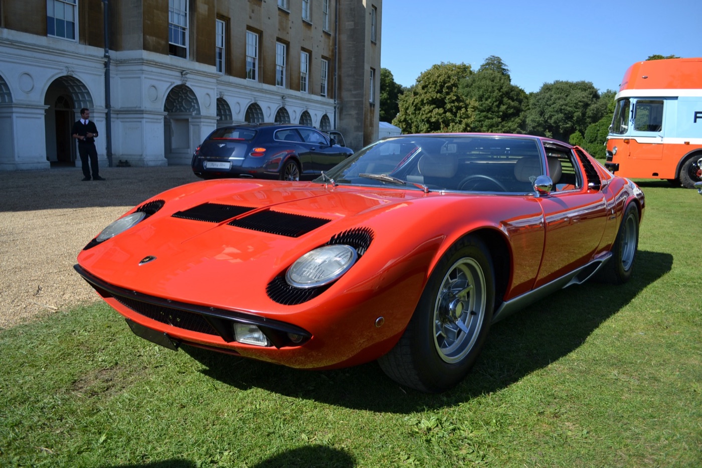 An orange-red Lamborghini Miura on the grass beside the stone colonnade of Syon House, a Gulf-liveried transporter behind