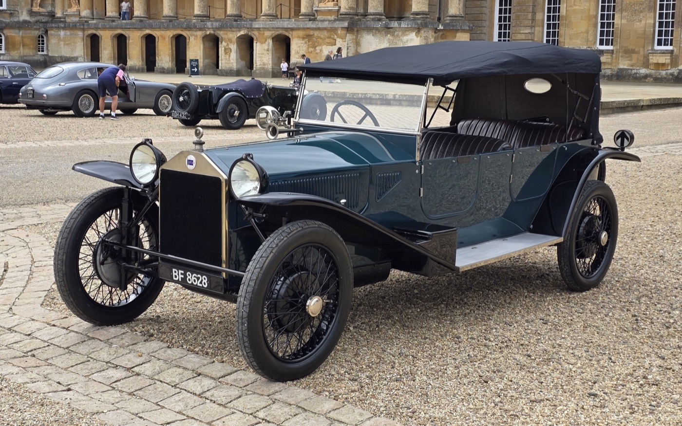 A dark green 1924 Lancia Lambda tourer with its hood up on the gravel of the Great Court of Blenheim Palace, an Aston Martin DB2 and a vintage sports car behind