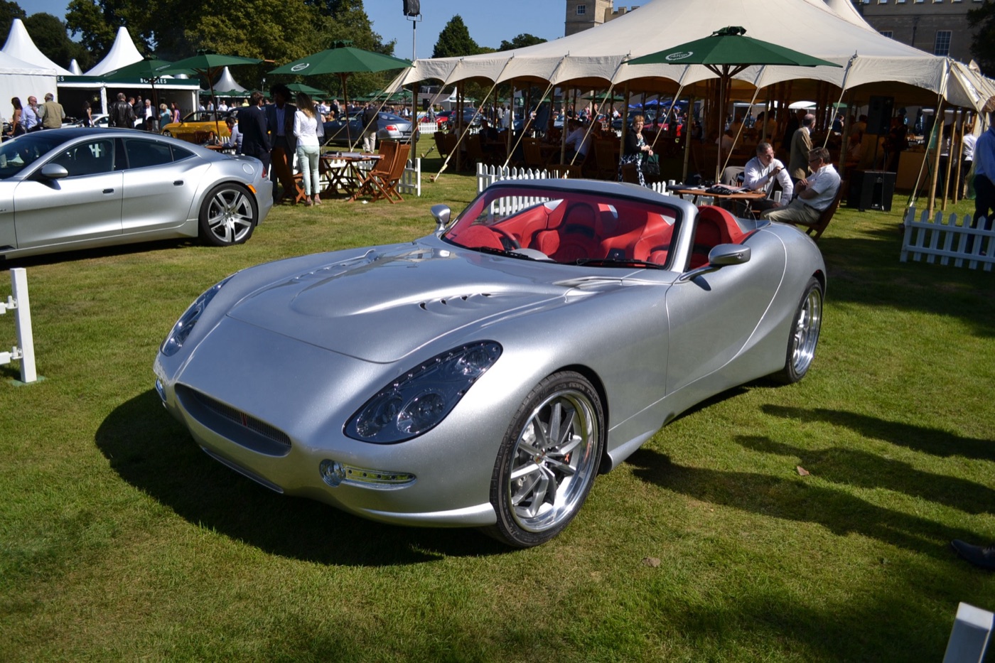 A silver Trident Iceni roadster with a red interior on the grass, with marquees, parasols and guests behind