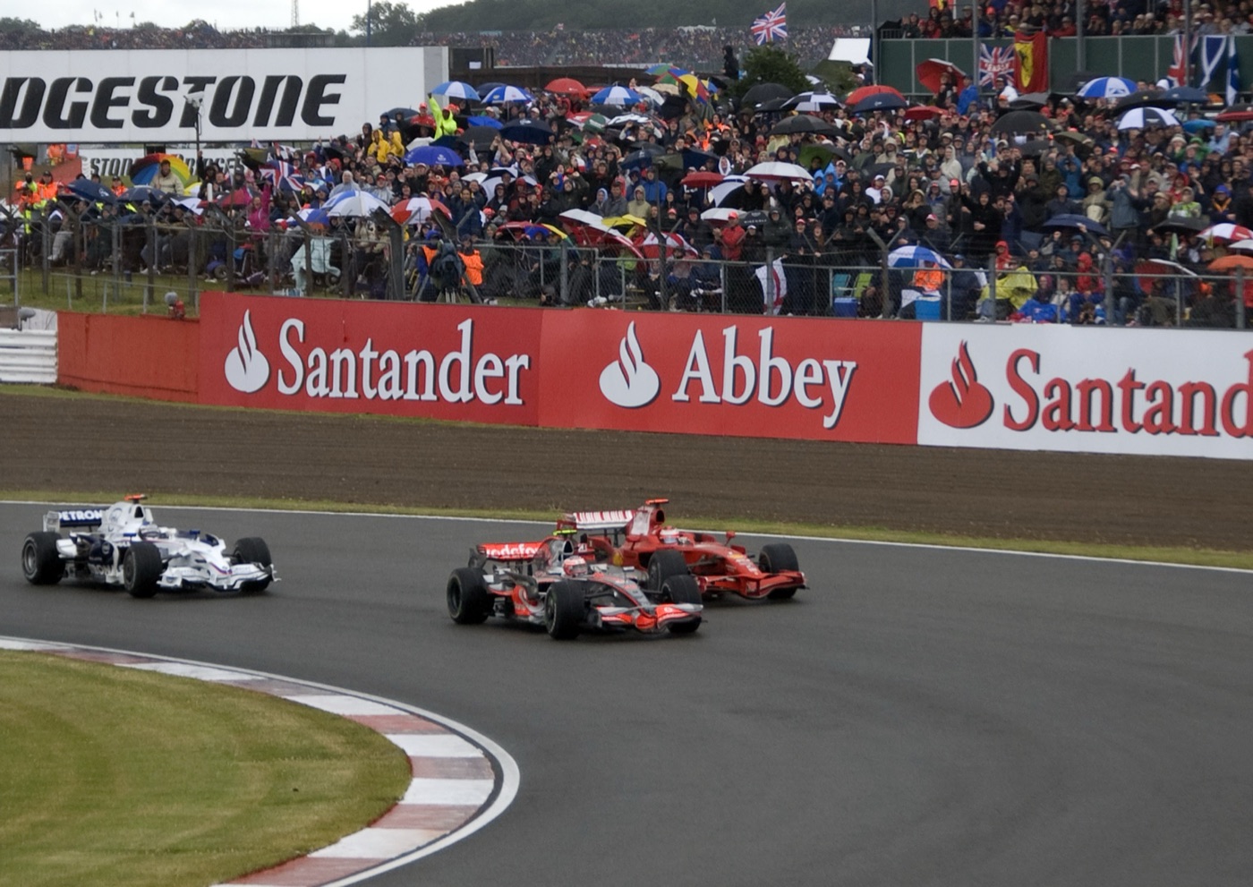 A silver McLaren, a red Ferrari and a white BMW Sauber Formula One car rounding a bend at Silverstone in the wet, in front of a crowd holding umbrellas