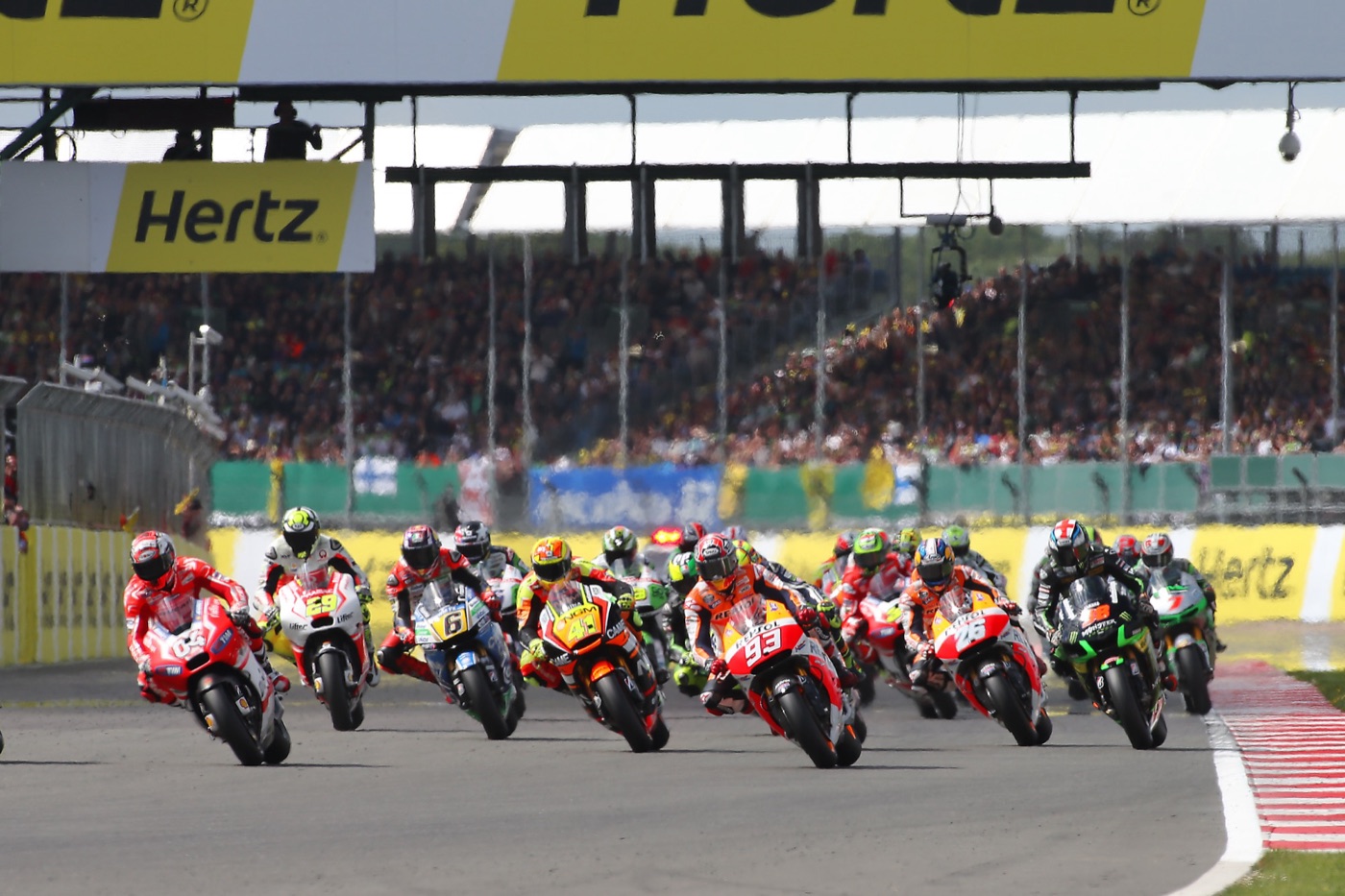 A pack of MotoGP riders on the opening lap at Silverstone, led by a red Ducati, with a full grandstand behind