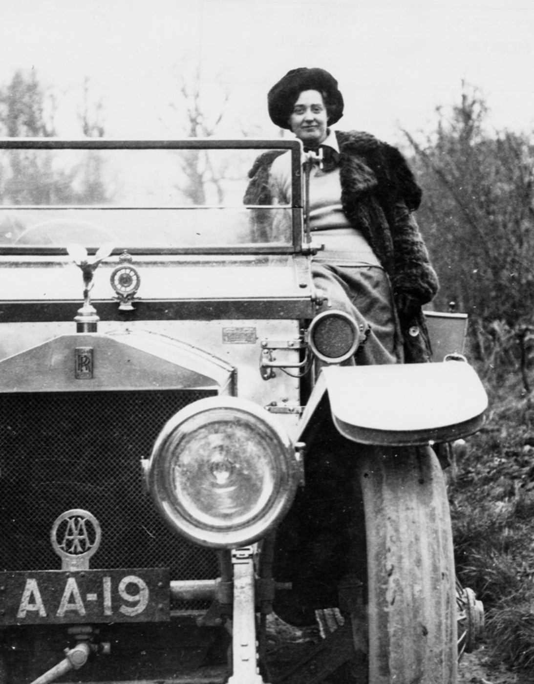 Black and white photograph of Eleanor Thornton in a fur coat and hat seated on an open Rolls-Royce Silver Ghost registered AA-19, with a mascot on the radiator