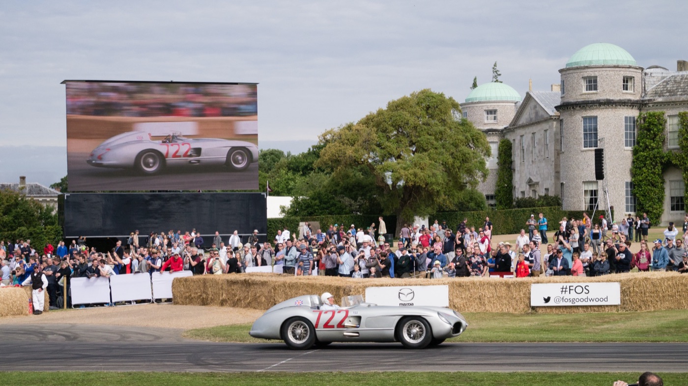 The silver Mercedes-Benz 300 SLR with red number 722 on its side drives past hay bales and a crowd at Goodwood House, with the same car shown on a large screen