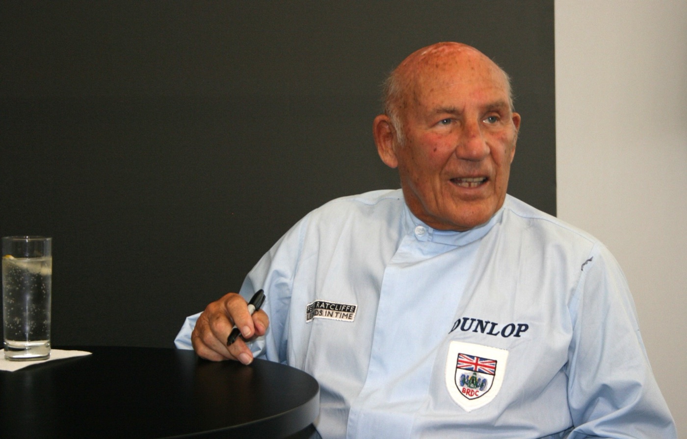 Sir Stirling Moss, in his eighties, in a pale blue shirt with Dunlop and BRDC badges, holds a marker pen at a table while speaking