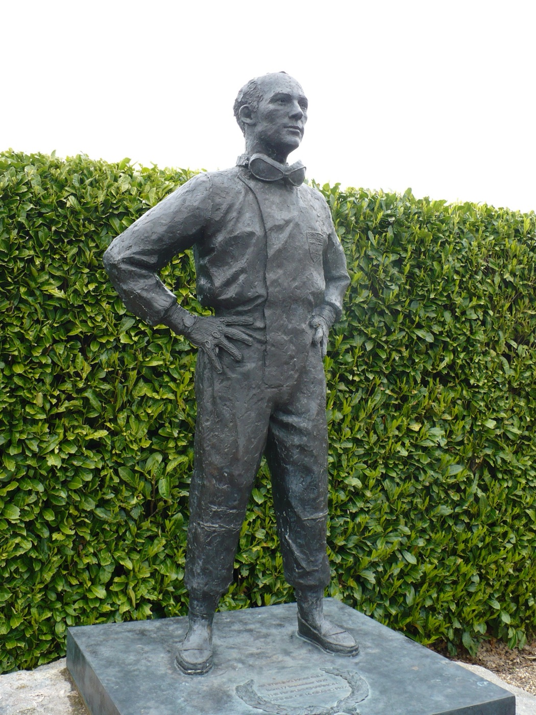 A bronze statue of Stirling Moss in racing overalls with goggles round his neck and hands on hips, in front of a laurel hedge