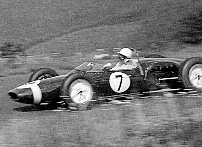 Stirling Moss in a white helmet drives the dark Rob Walker Lotus numbered 7 at speed, the background blurred