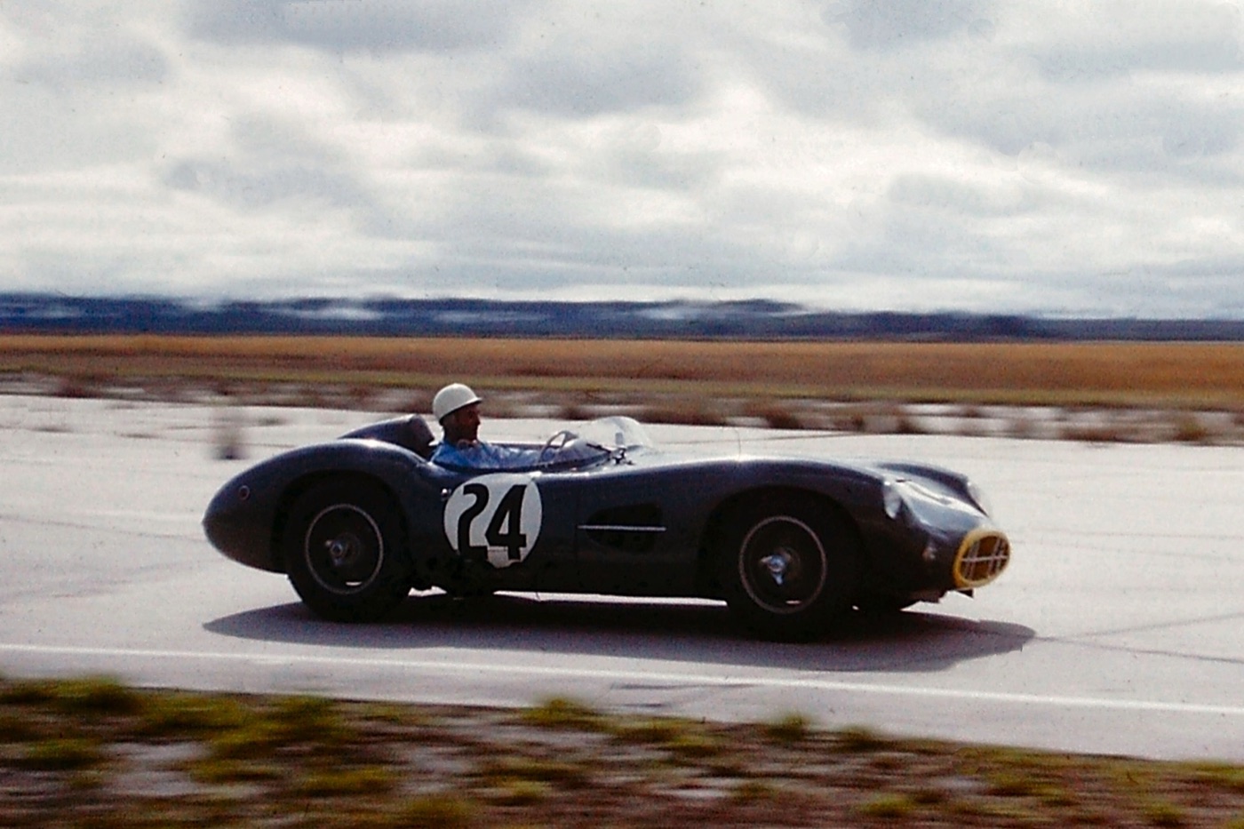 Stirling Moss in a white helmet drives a dark green Aston Martin DBR1 numbered 24 along a wide airfield circuit under a cloudy sky
