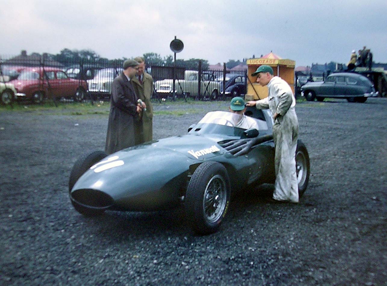 A dark green Vanwall Formula One car on gravel in the Aintree paddock, a driver in the cockpit and a mechanic in white overalls beside it, with parked cars behind