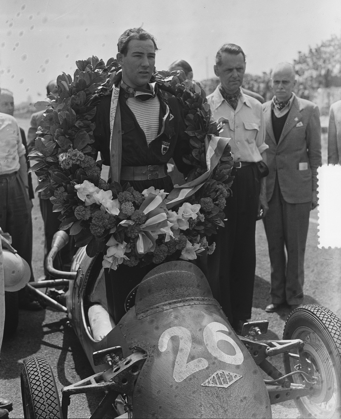 A young Stirling Moss in overalls and goggles, a large laurel wreath over his shoulders, standing in a Kieft 500 cc racing car numbered 26
