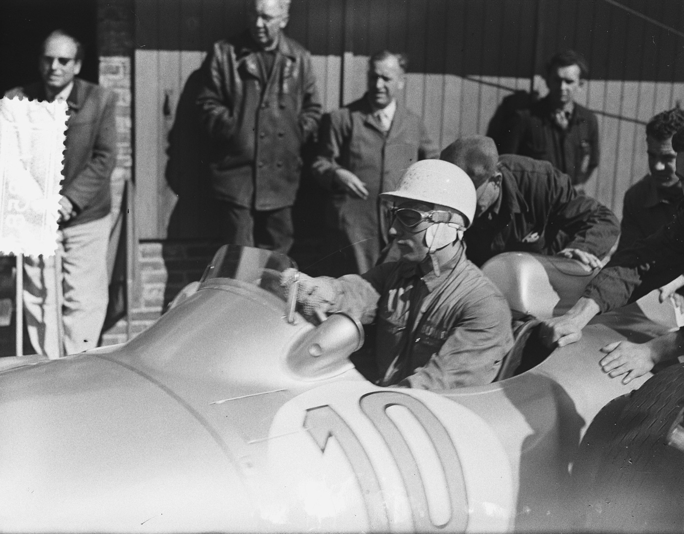 Stirling Moss in a white helmet and goggles sits in the silver Mercedes-Benz W196 numbered 10 while mechanics push the car in the pits