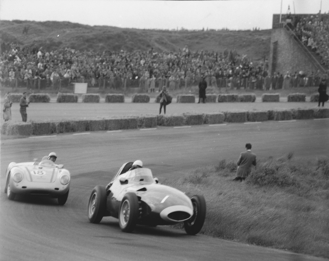 A Vanwall Formula One car leans through a sweeping corner at Zandvoort, followed by a silver Porsche, with packed grandstands and dunes behind