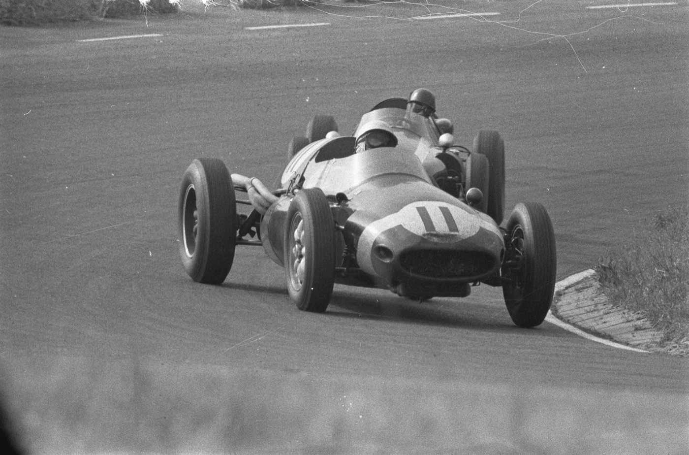 A rear-engined Cooper Formula One car numbered 11 rounds a corner in front of another single-seater on a grey track