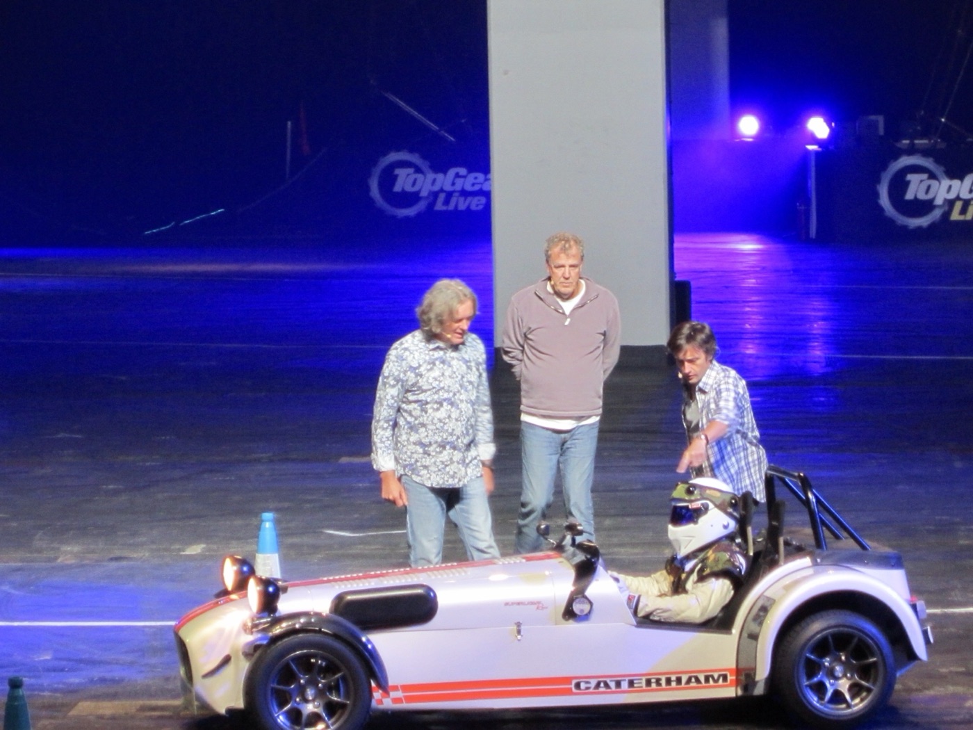The Stig in a white helmet at the wheel of a Caterham Seven, with James May, Jeremy Clarkson and Richard Hammond beside the car