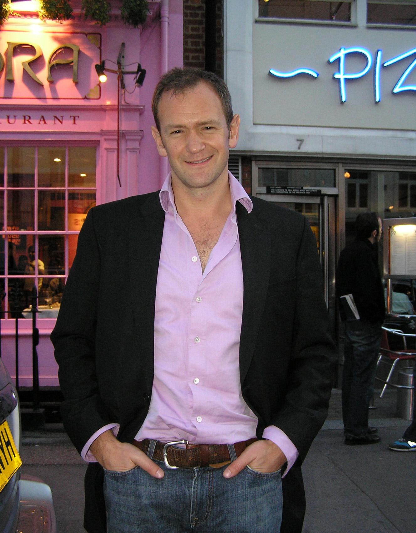 Alexander Armstrong in a dark jacket and pink shirt standing on a London street at dusk