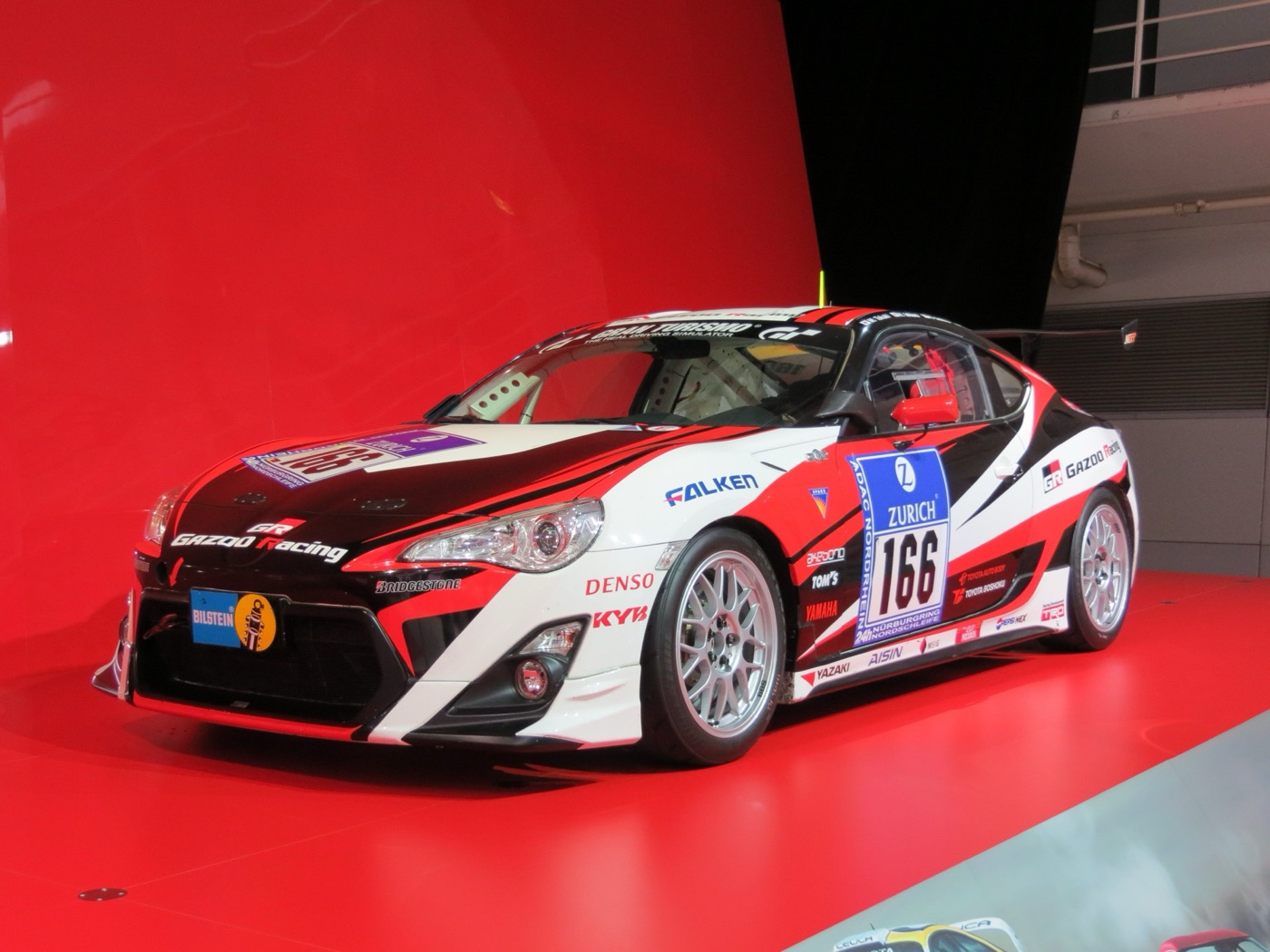 White, black and red Gazoo Racing Toyota 86 race car number 166 with Nürburgring 24h stickers on a red stand