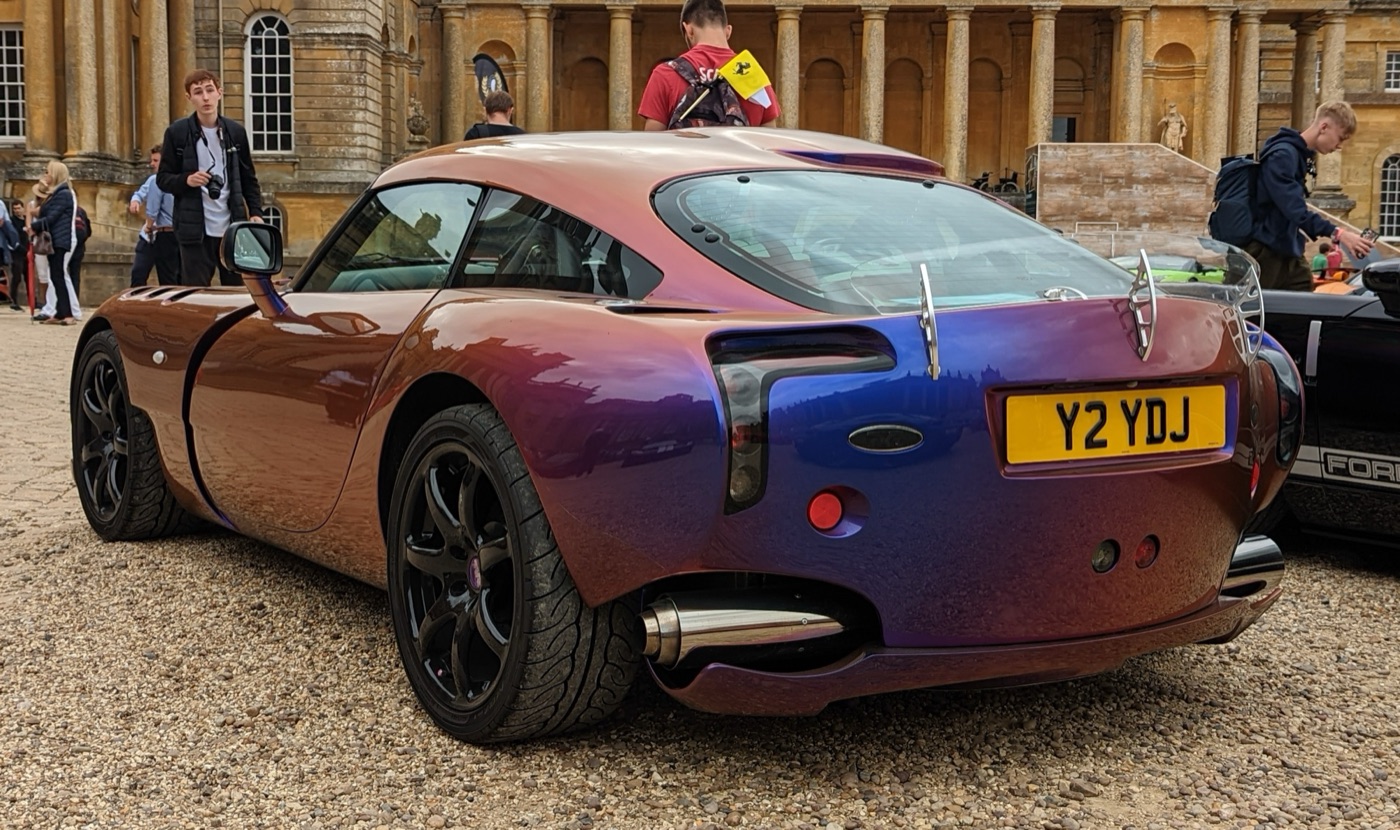 Rear view of a colour-shift purple and bronze TVR Sagaris with a clear rear spoiler at a car show in front of a stately home
