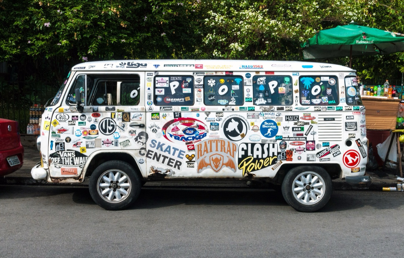 White Volkswagen T2 Kombi covered in skate and surf stickers, parked by a pavement and used as a street stall