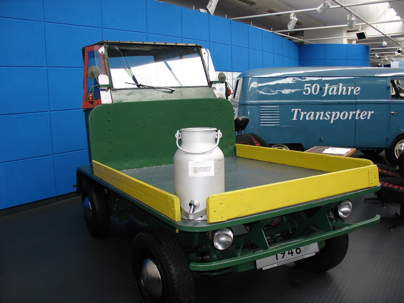 Green Volkswagen Plattenwagen factory parts carrier with a flat yellow-edged load bed and a milk churn on board, beside a blue T1 panel van