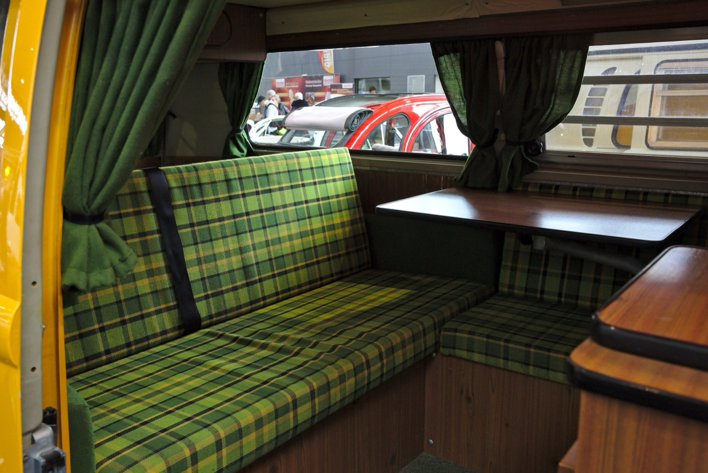 Interior of a Volkswagen T1 Westfalia camper with green and yellow tartan bench seats, a wooden table, cupboards and green curtains