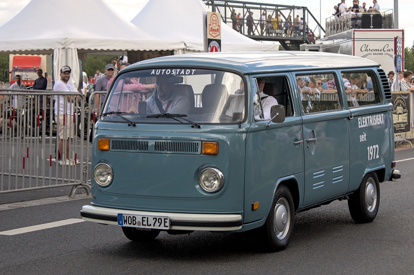 Grey-blue Volkswagen T2 bay-window bus with ELEKTRISIERT SEIT 1972 lettering on its side, driving past a crowd at a classic car event