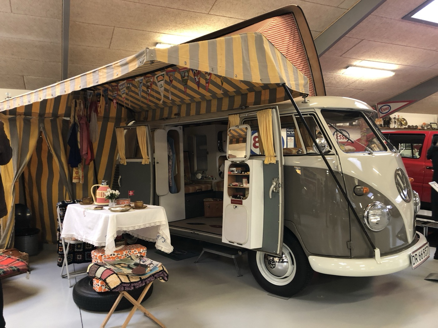 Grey and white 1963 Volkswagen T1 Westfalia SO 36 camper with its doors open, a striped awning, a table laid with a cloth and camping gear, in a museum