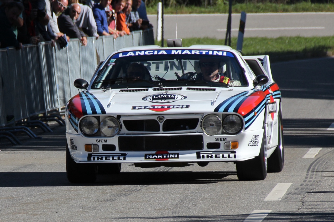 Lancia 037 Rally in white Martini colours with four spotlights, driving towards the camera past a crowd behind barriers