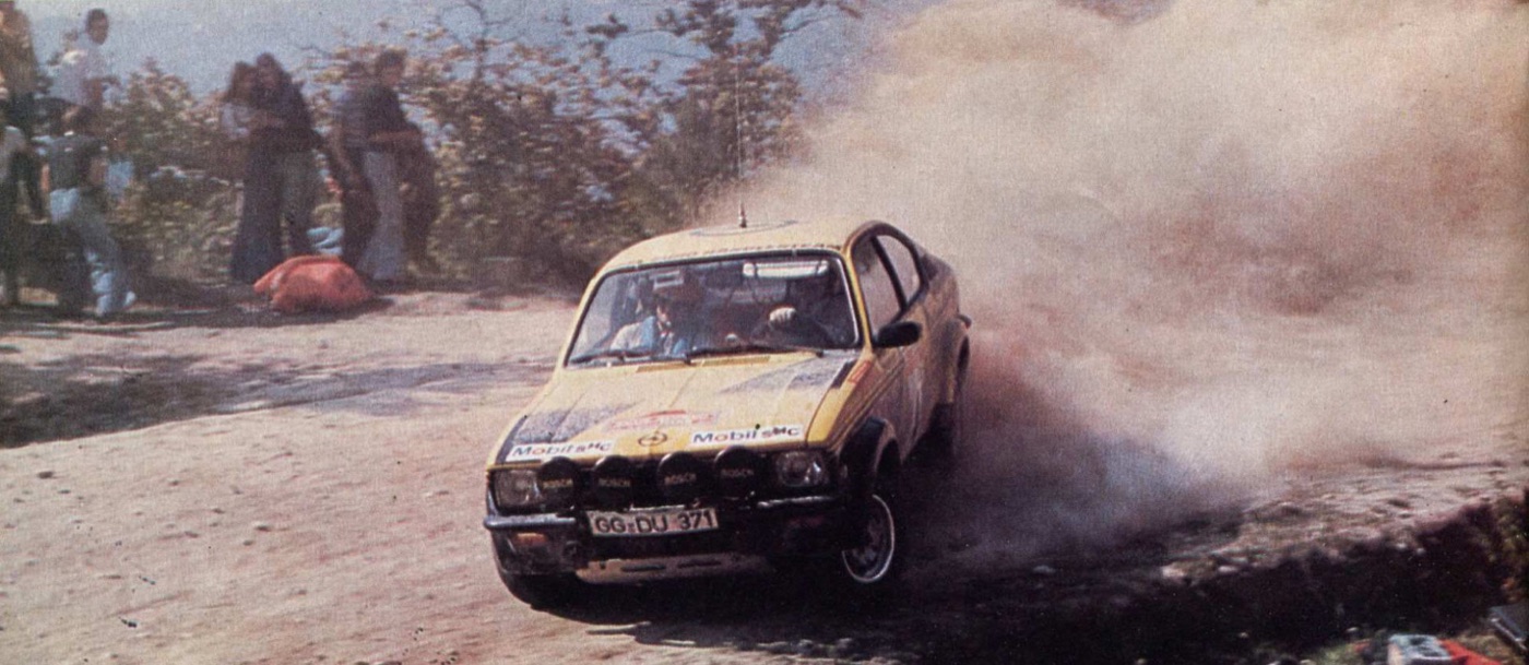 Walter Röhrl’s yellow and black Opel Kadett GT/E sliding through dust on a gravel stage, spectators behind