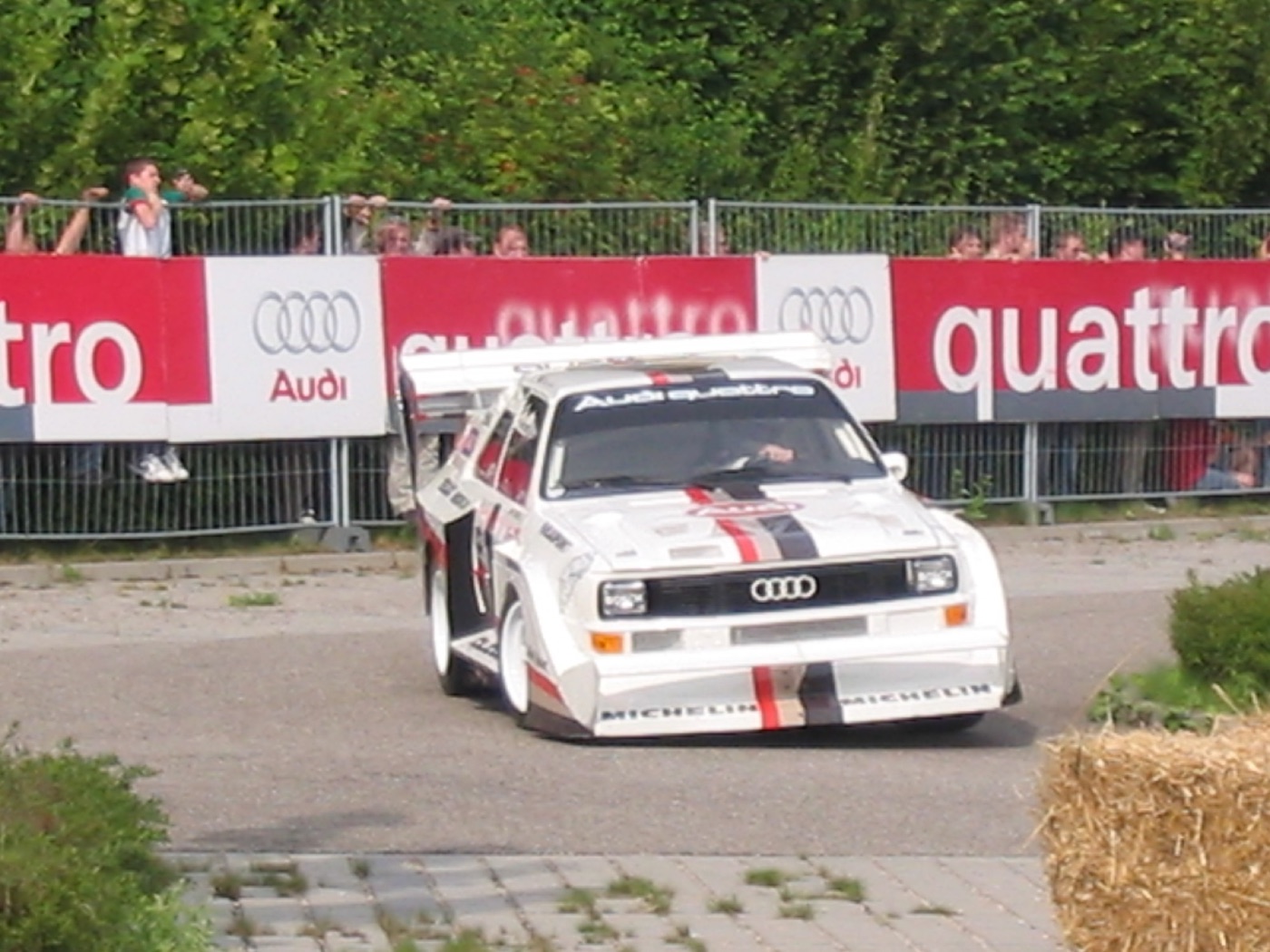 White Audi Sport Quattro S1 Pikes Peak car with a huge rear wing and front splitter, cornering past Audi quattro banners