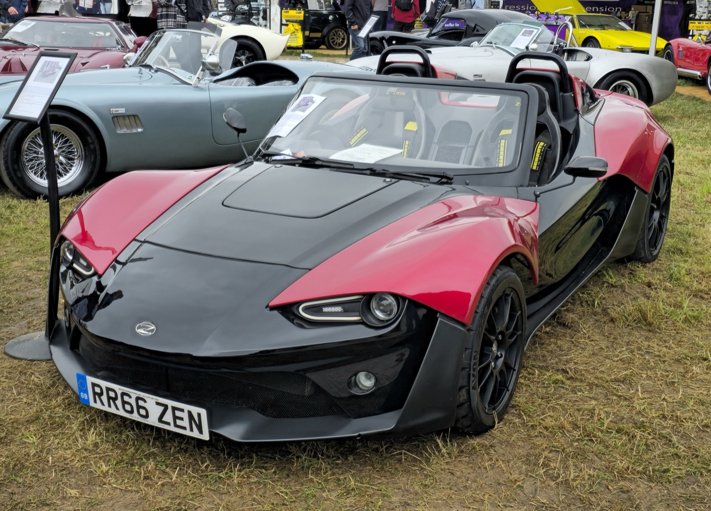 Red and black Zenos E10 with a windscreen and twin roll hoops, parked on grass among other sports cars at a show